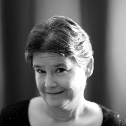 A greyscale photograph of the author, a middle aged white woman with brown hair and eyes, smiling out at the viewer.